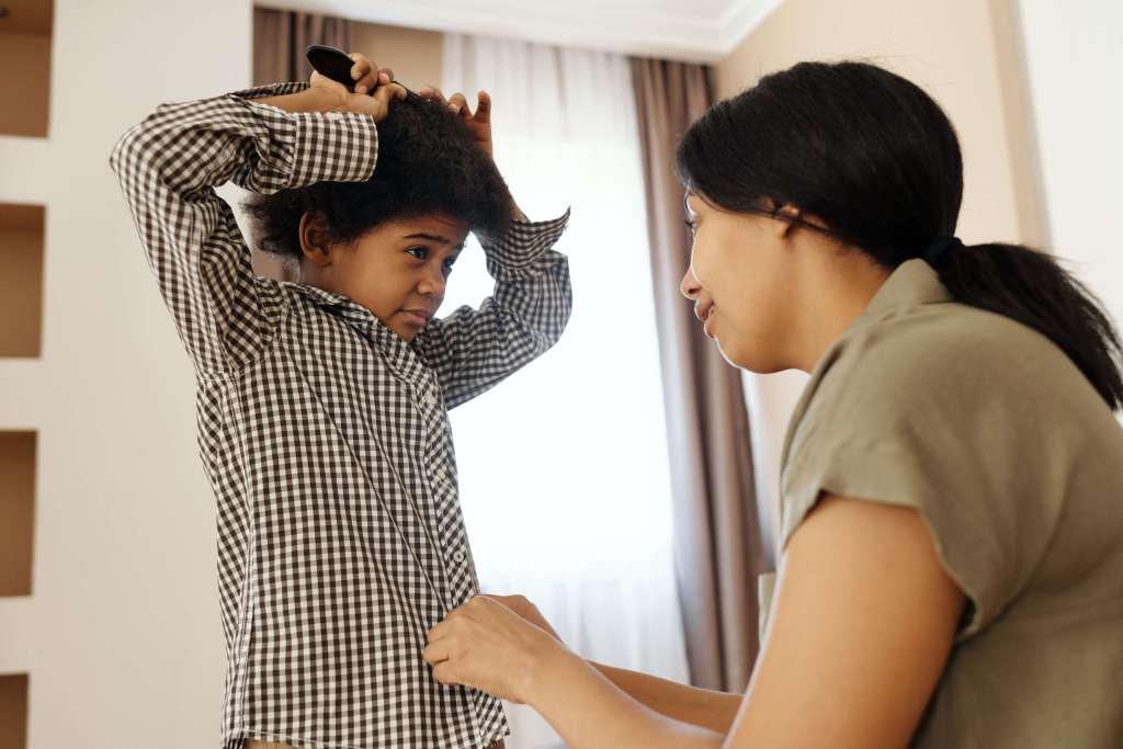Image of mother buttoning her son's shirt while he brushes his hair. Montessori Preschool